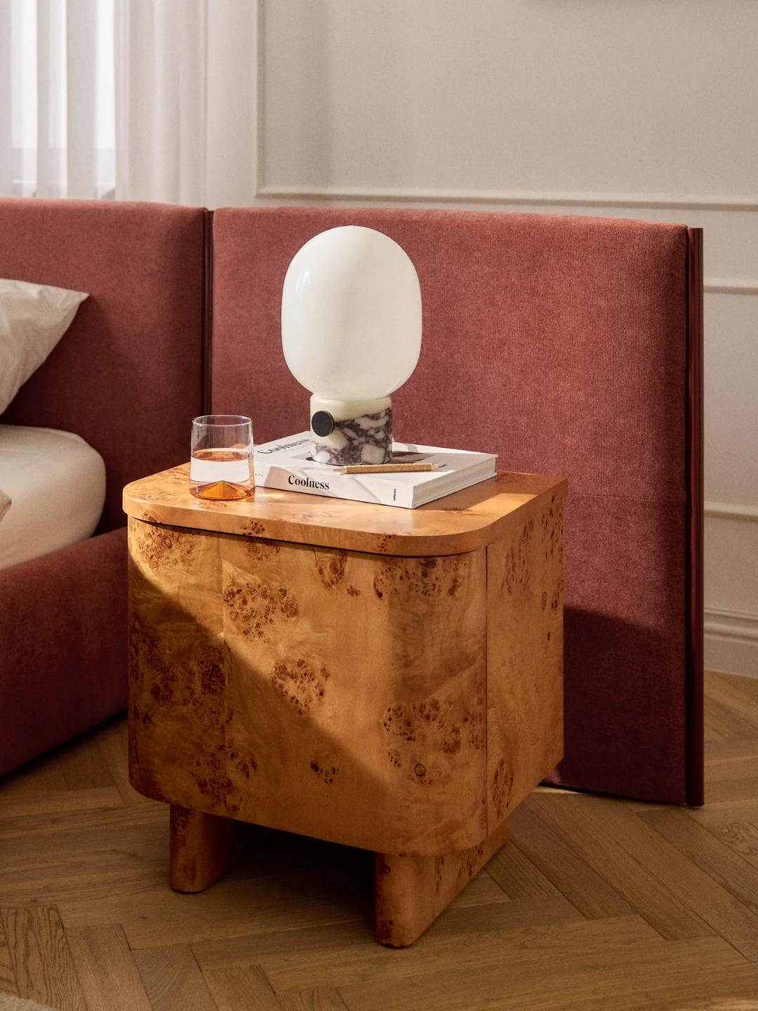 A bedroom with a curved burl nightstand by an extended headboard