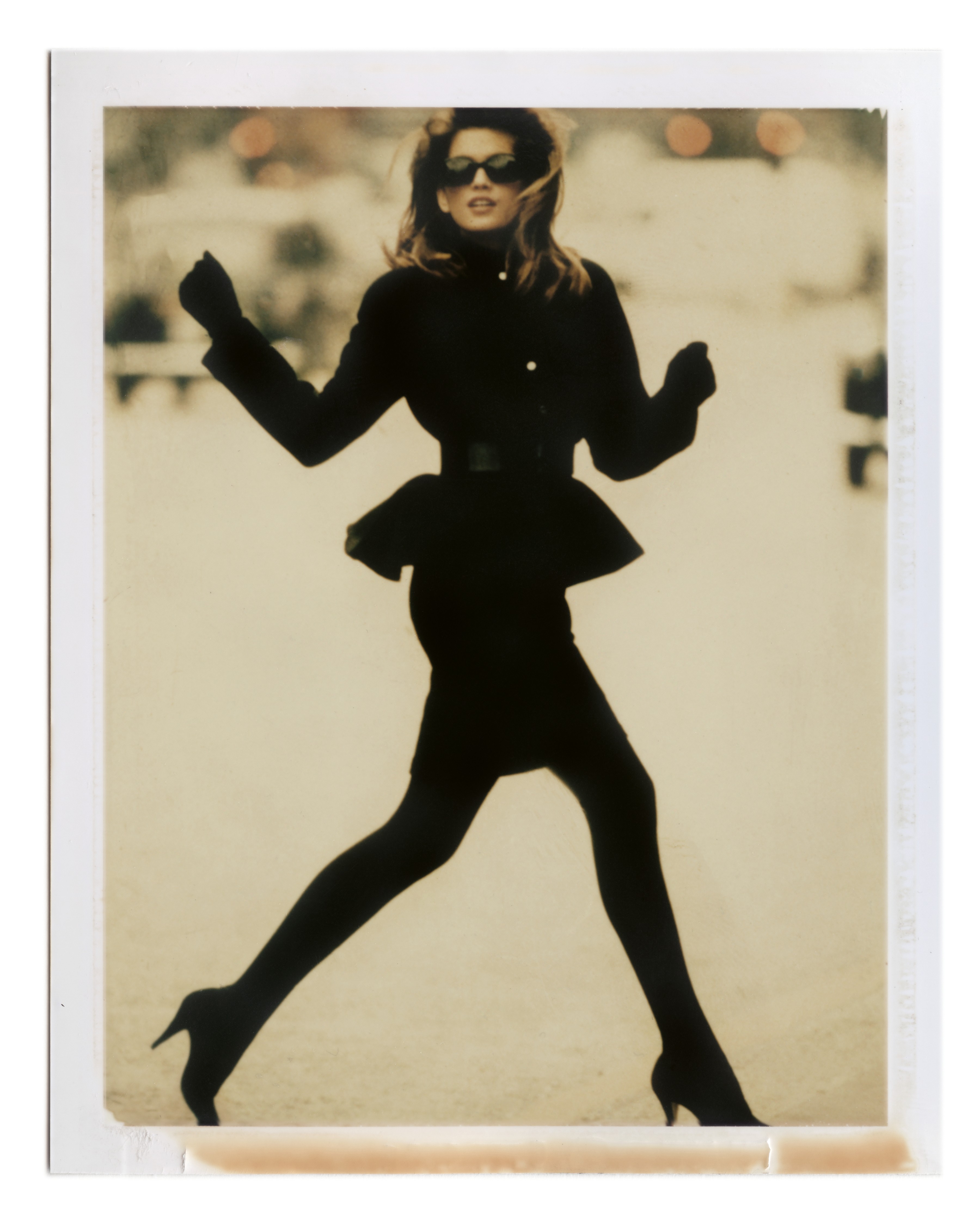 A model in a black belted jacket and skirt leaps with arms outstretched and sunglasses on, captured in a faded vintage Polaroid.
