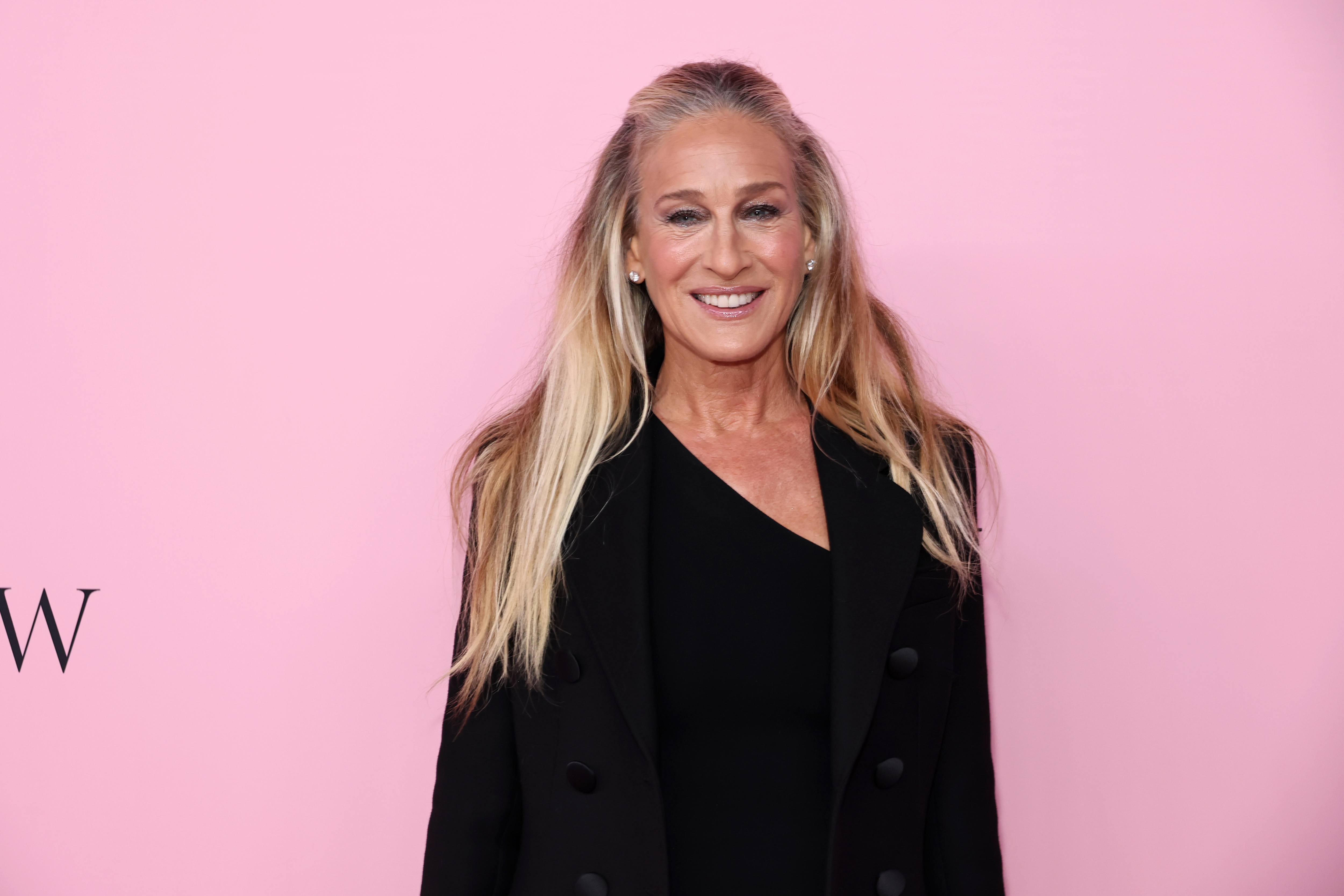 a woman with long blonde hair and dark makeup standing in front of a pink wall with a black jacket on