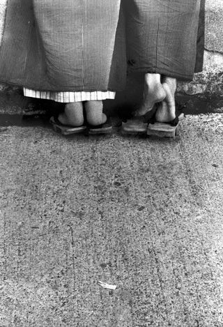 Two Japanese people's feet are captured in traditional sandals and kimonos in black and white from behind.