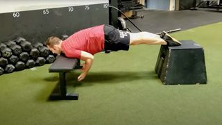 YouTube: Man performing a prone Chinese plank with fronts of shoulders rested on an exercise bench and feet rested on a box behind him