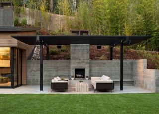 Sloped backyard ideas featuring a concrete seating area with black pergola and outdoor fireplace.