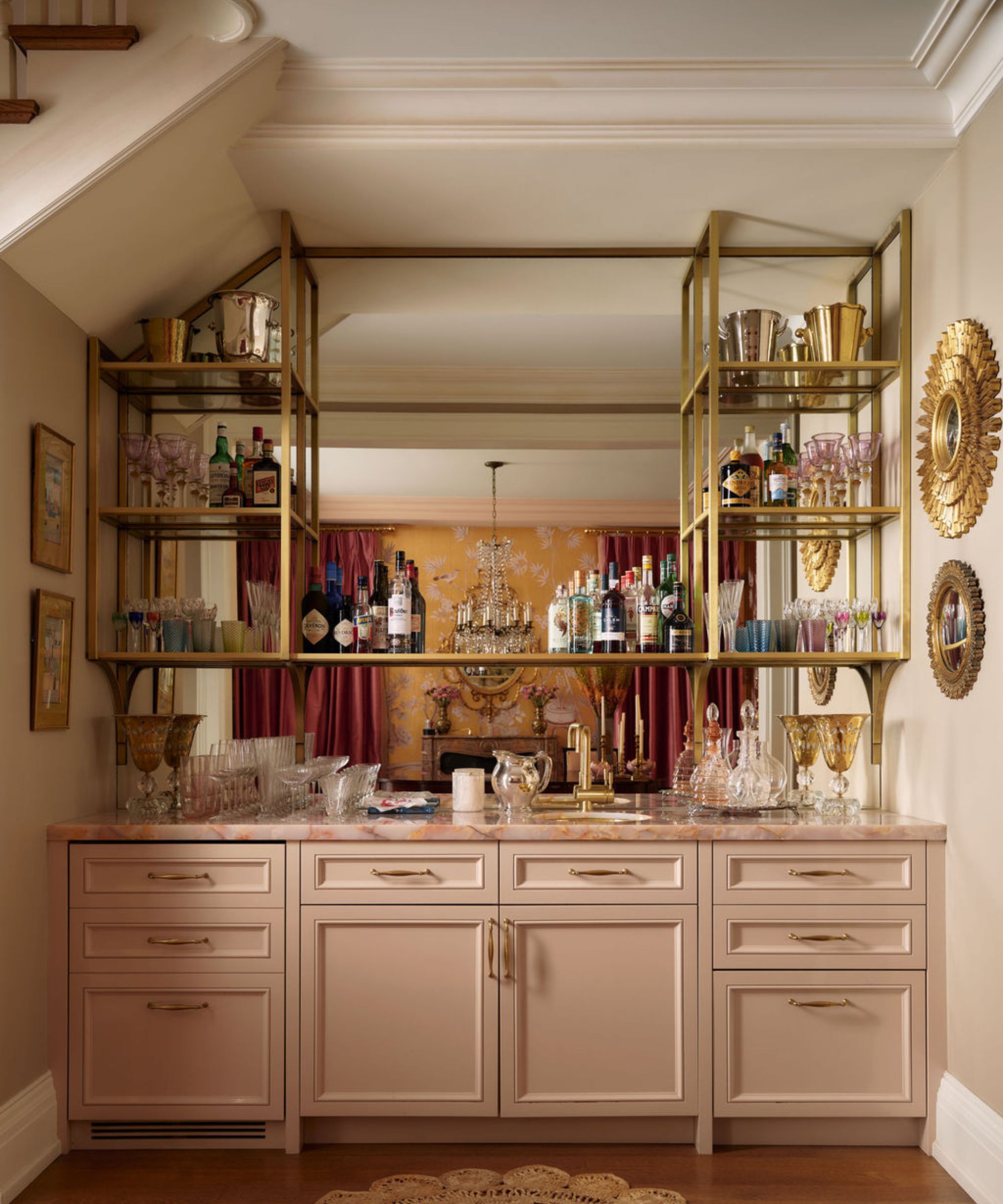 a pink home bar tucked under a staircase with brass and glass open shelving over a mirrored wall reflecting the dining room