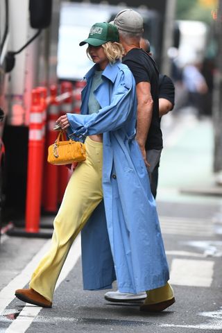 Gigi Hadid wearing a blue trench coat, butter yellow trousers, Birkenstocks, and a yellow Miu Miu bag in New York City July 2024