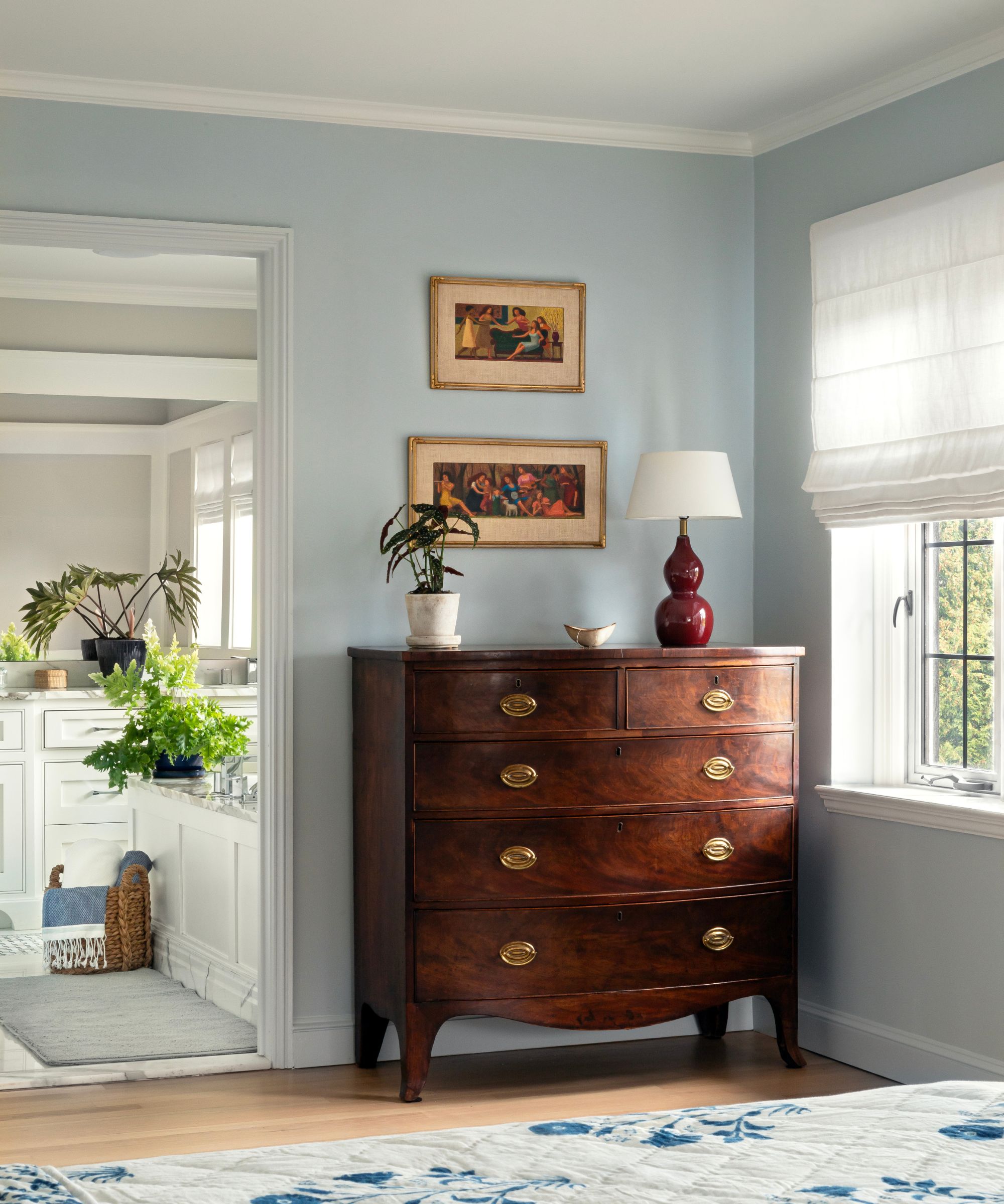 Pale blue bedroom with a dark mahogany dresser in the corner with a red lamp and potted plant on top, and two antique paintings above. Beside it is a window with a white Roman blind.
