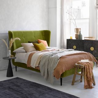 how to make a guest room look more expensive, white bedroom with green velvet upholstered bed, sideboard, white floorboards, grey rug, side table, stool, ochre throw