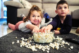 A young sister and older brother watching a film whilst eating popcorn out of a bowl. They are lying on a rug in the living room.