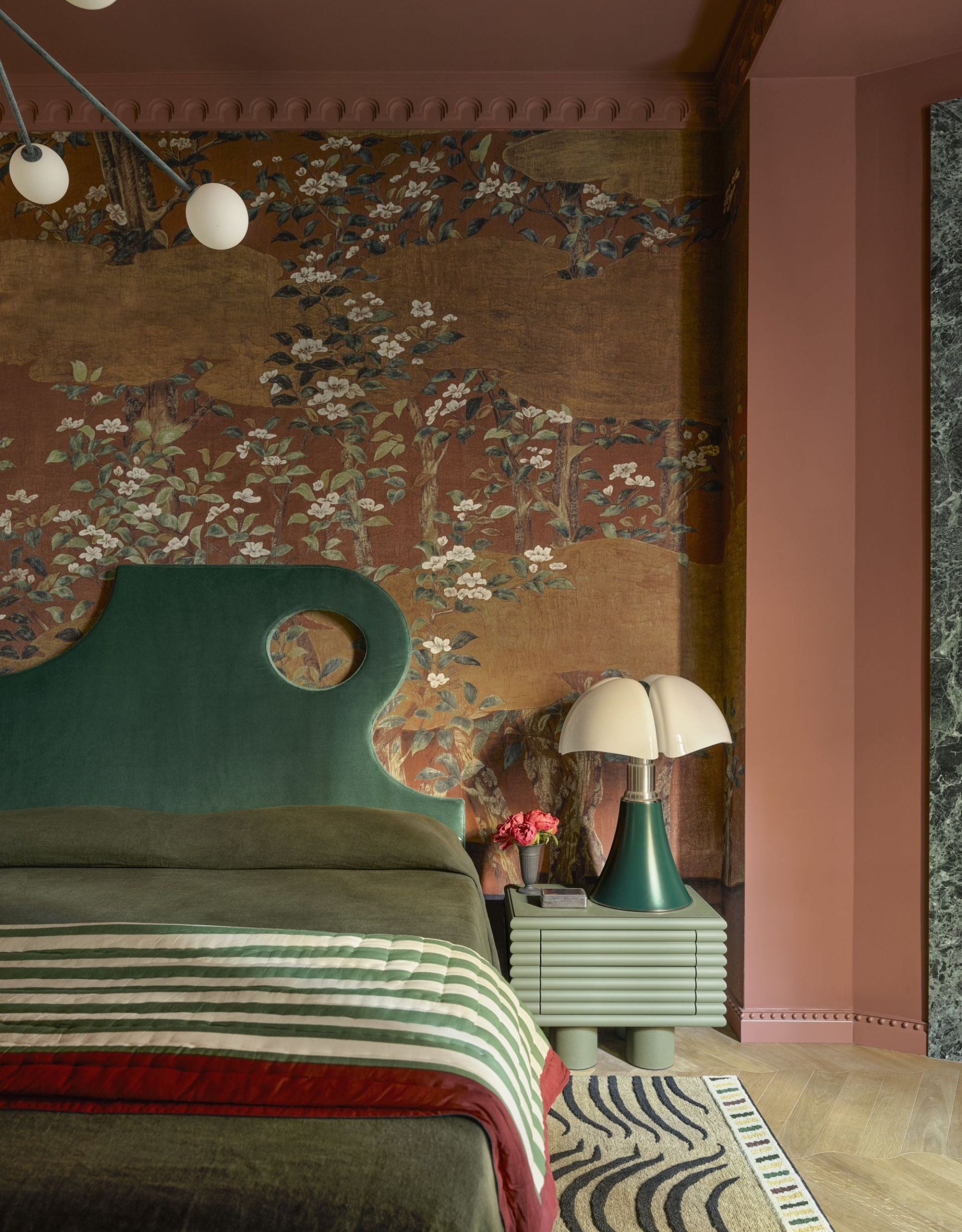 A bold modern bedroom decorated with deep earth tones with patterned wallpaper, an abstract green headboard and green bedding and bedside with an abstract green bedside lamp