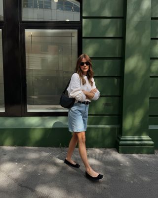 British female fashion influencer Kim Turkington poses on a London sidewalk wearing black Ray Ban sunglasses, a white crewneck sweater, black sling shoulder bag, black belt, long denim shorts, and black square-toe ballet flats