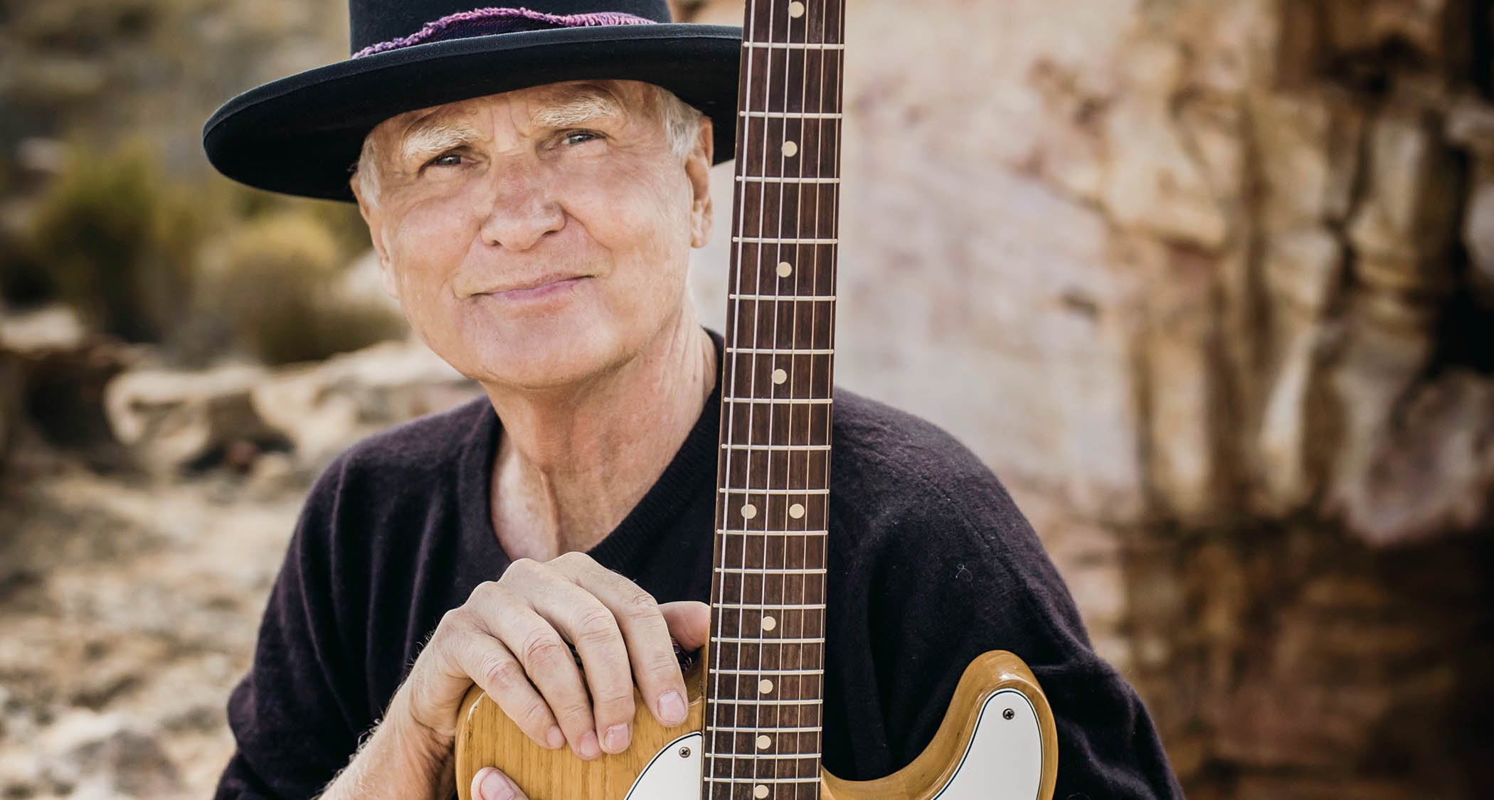 Steve Louw is picture in a wide-brimmed hat with his guitar in the desert.