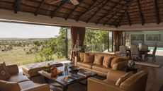 Image of a living room with lots of brown and natural decor. There is a thatched roof and the walls are open to views of the Serengeti.