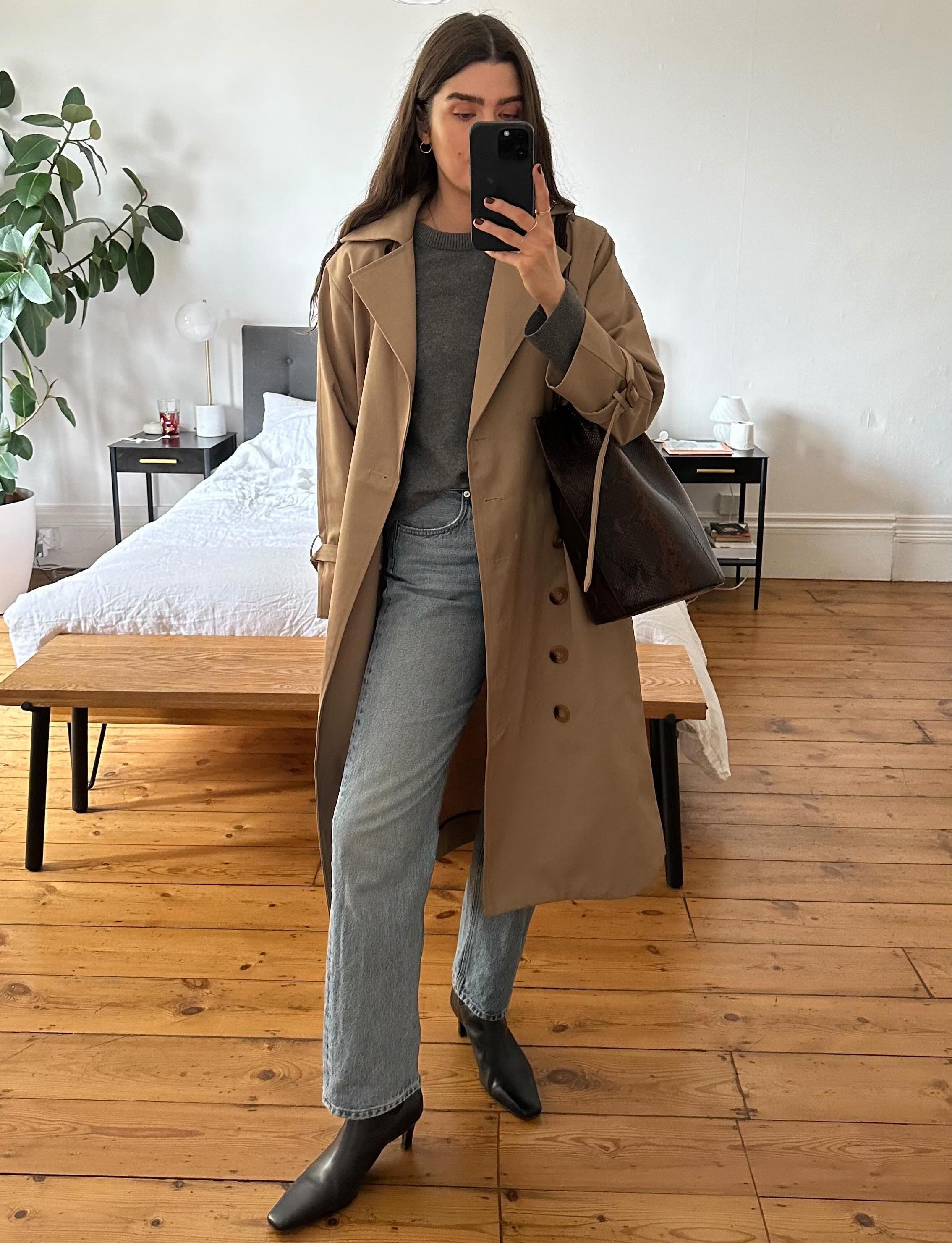 British fashion influencer Anna Howard poses for a mirror selfie wearing a trench coat, gray crewneck sweater, straight-leg jeans, leather tote bag, and black square-toe boots