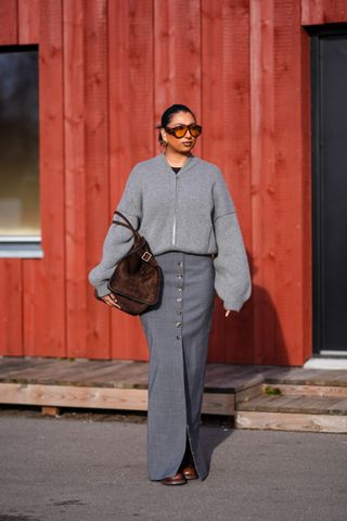 A guest wears sunglasses, earrings, a gray wool oversized cardigan, a brown suede bag, a gray long slit skirt , brown leather boots , outside Gestuz, during the Copenhagen Fashion Week AW24 on February 01, 2024 in Copenhagen, Denmark.