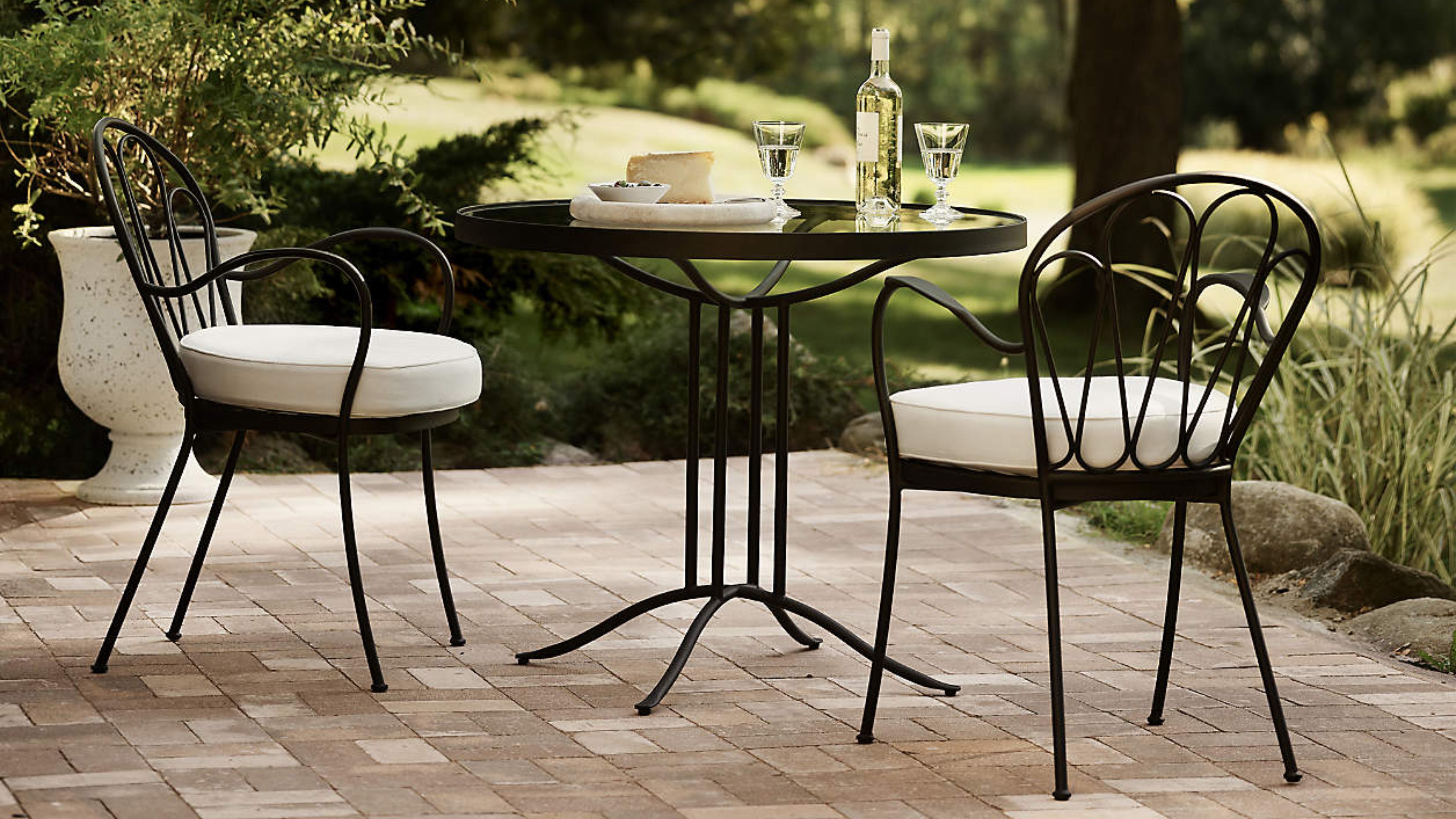 Whimsical wrought iron bistro table set topped with white wine and stemware, set atop of a brick patio overlooking a lush backyard
