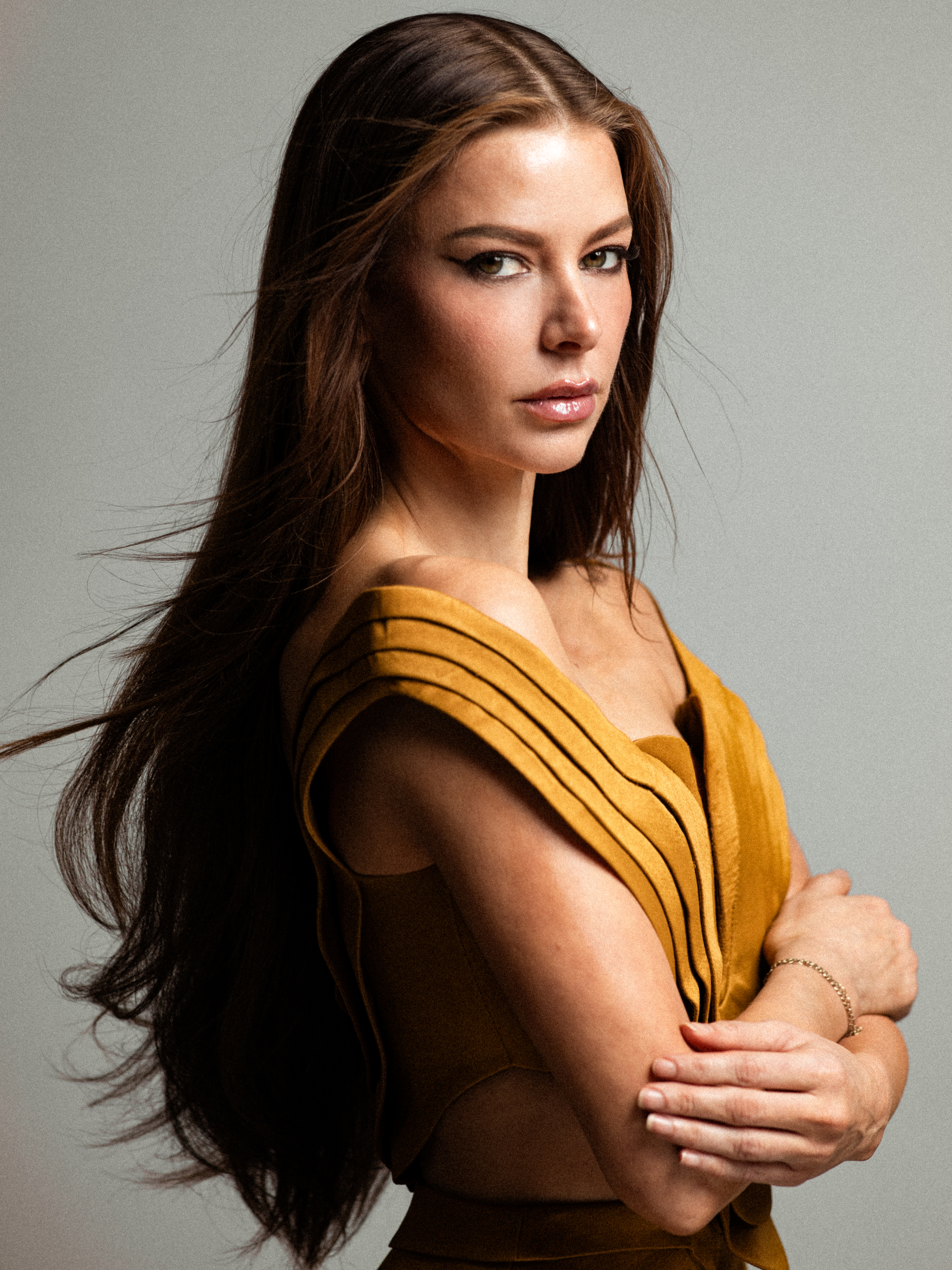 ariana madix wears a gold layered short dress and her brown hair down long and windswept as she poses crossing her arms and looking over her shoulder