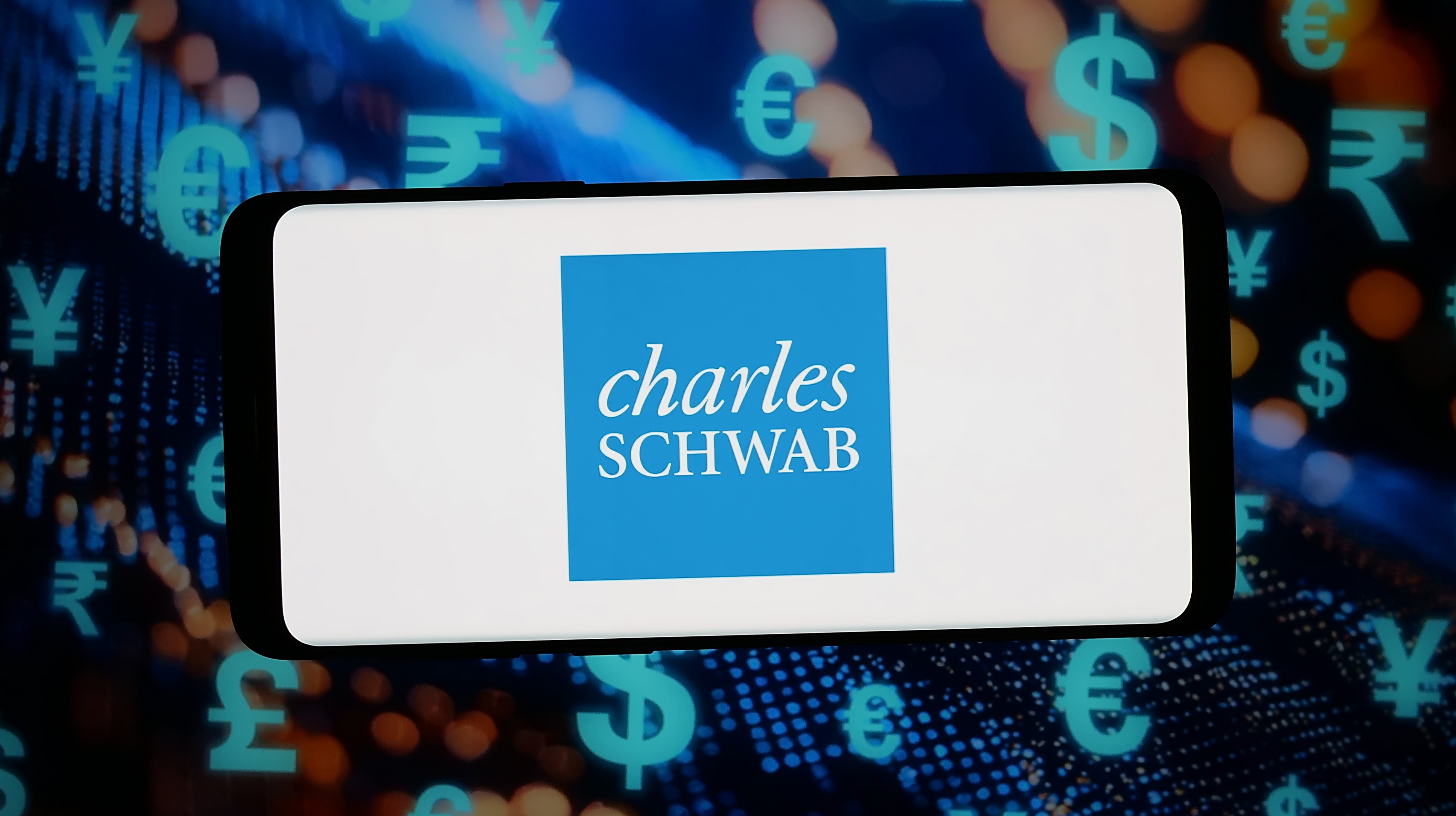 The Charles Schwab logo displayed on a smartphone with various currency symbols in the background.