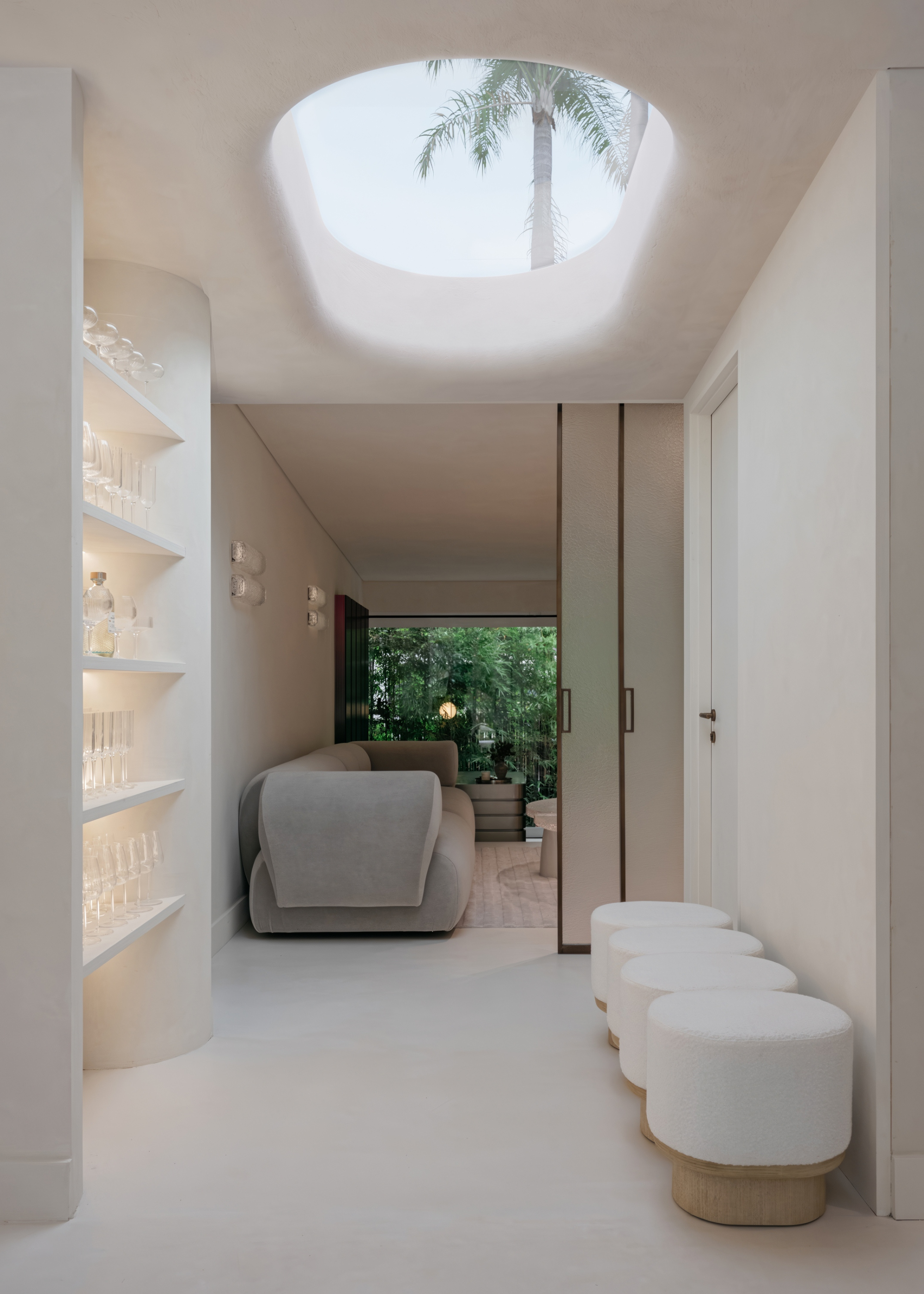 a neutral hallway with shelving storage with glasses with stools facing it leading and a skylight leading into a living area