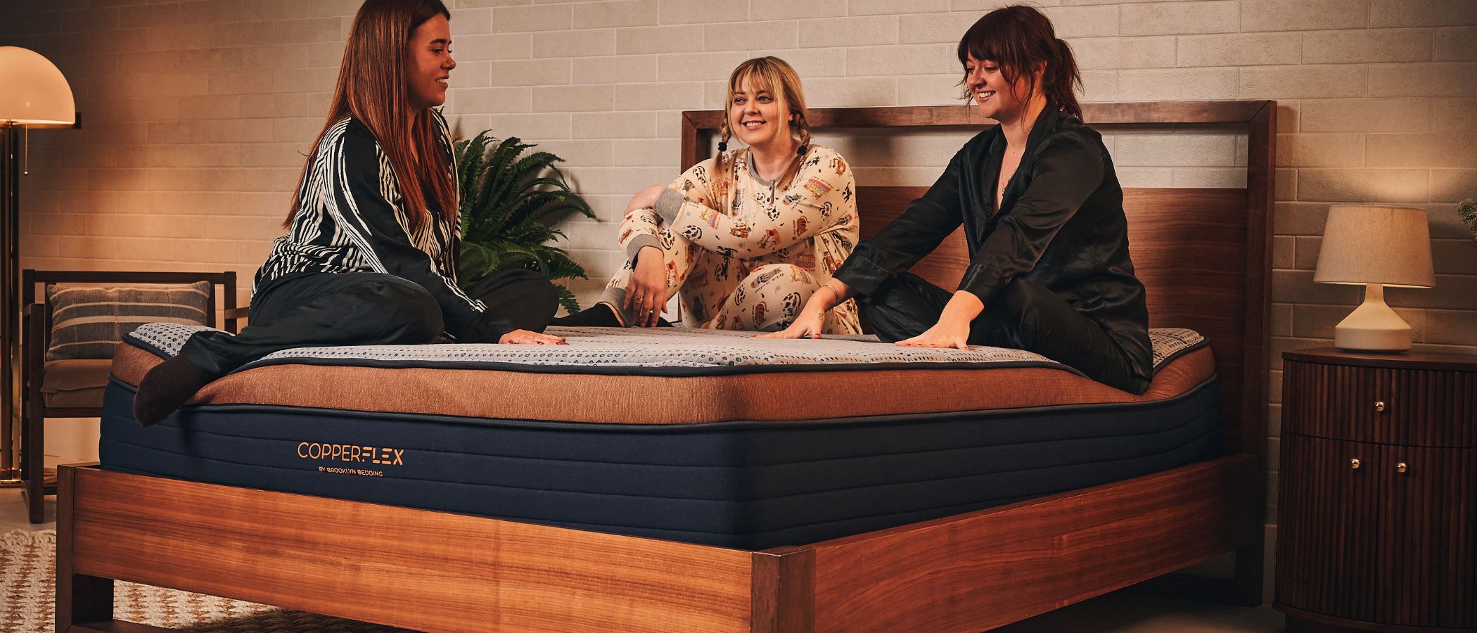Three women in pyjamas sit cross-legged on a Brooklyn Bedding CopperFlex mattress, chatting and smiling. The mattress features a textured blue-grey top, a copper-colored border, and a dark blue base with the Copperflex logo visible on the side. The bed has a modern wooden frame and headboard, set in a cozy bedroom with warm lighting. 