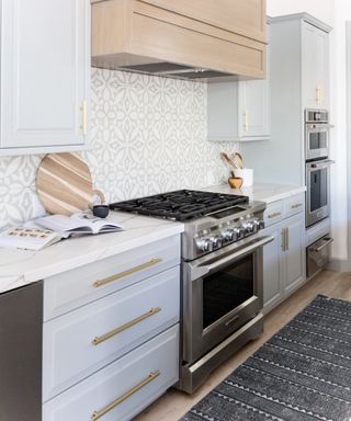 pale gray kitchen cabinets, pale gray patterned tiles backsplash, with dark gray rug, white walls, brass handles,