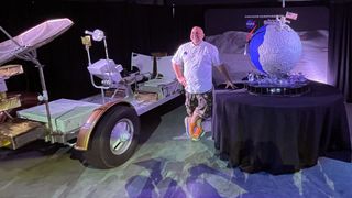 a smiling man in a white t shirt stands on a set next to a fake moon buggy and a cake that looks like the moon