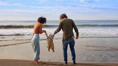 A mom and dad lift their toddler between them as they walk on the beach. 