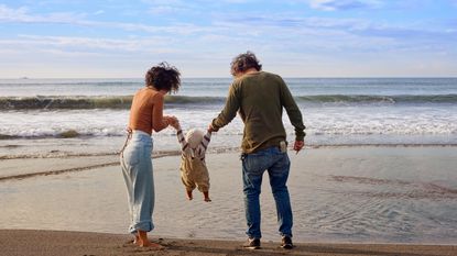 A mom and dad lift their toddler between them as they walk on the beach. 