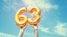 Gold foil number 63 balloons being held high in the air against a blue sky with clouds