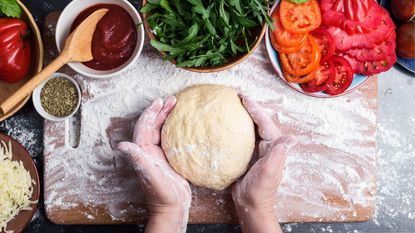 Hands holding a ball of pizza dough on a floured board with tomatoes, cheese and rocket in bowls around the edge