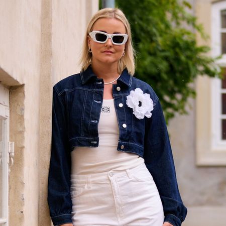 a woman wears a denim jacket with a white flower, white loewe tank top, white jeans, and white sunglasses.