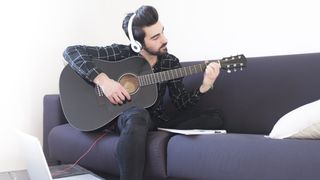 Young man at home playing guitar and wearing headphones connected to laptop