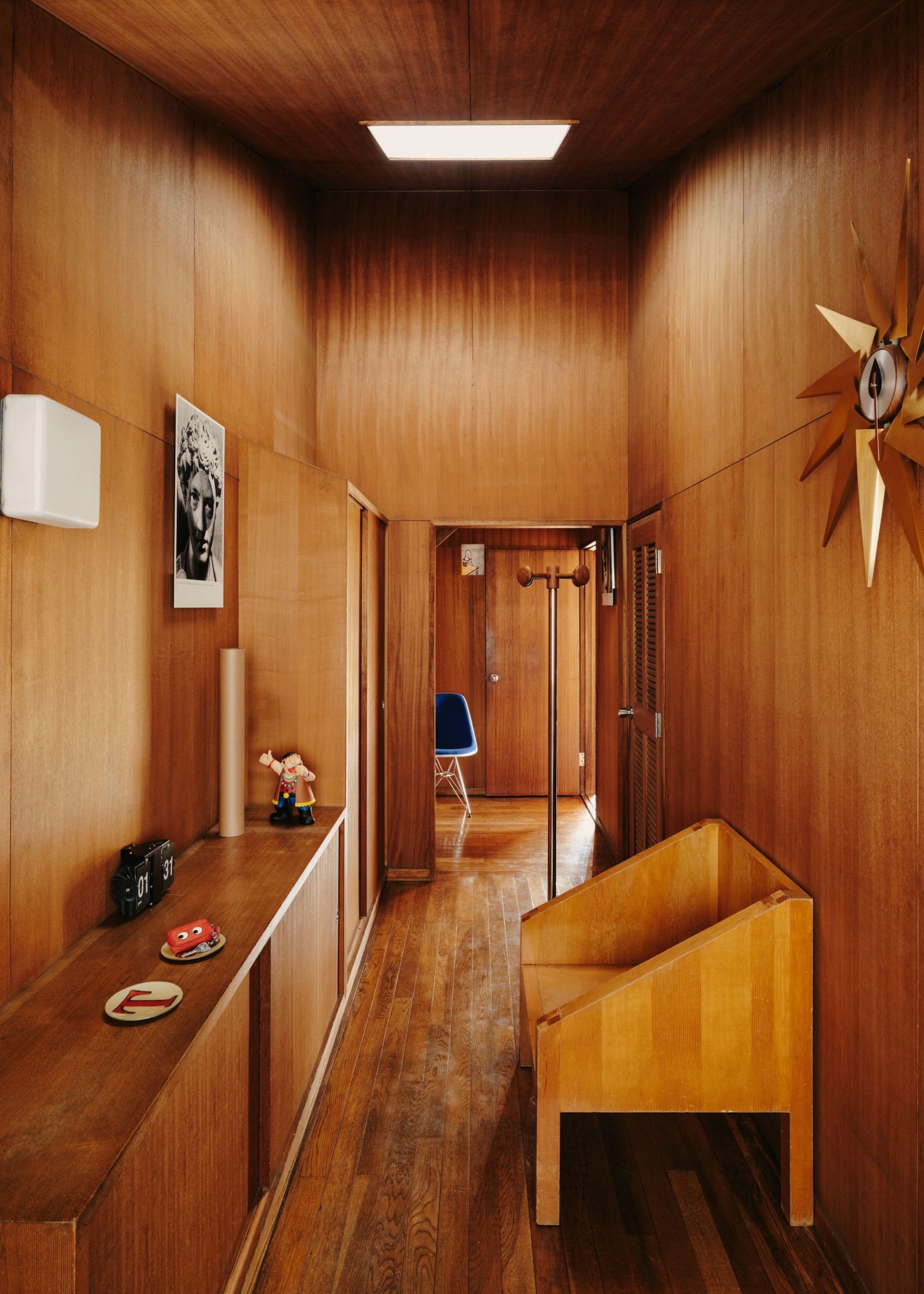A Japanese home with a wood drenched hallway with a skylight above, a wooden bench, and a console with sliding wood doors with minimal abstract decor on top