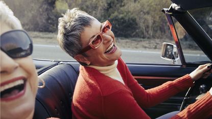 Two older women laugh as one drives a convertible.