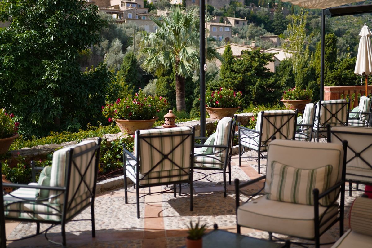 Outdoor terrace with cushioned chairs, potted flowers and palm trees overlooking a lush hillside.