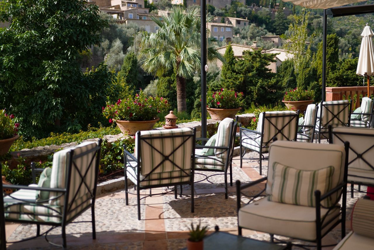 Outdoor terrace with cushioned chairs, potted flowers and palm trees overlooking a lush hillside.