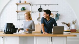 A man and woman in the kitchen using an air fryer, with the man on a laptop