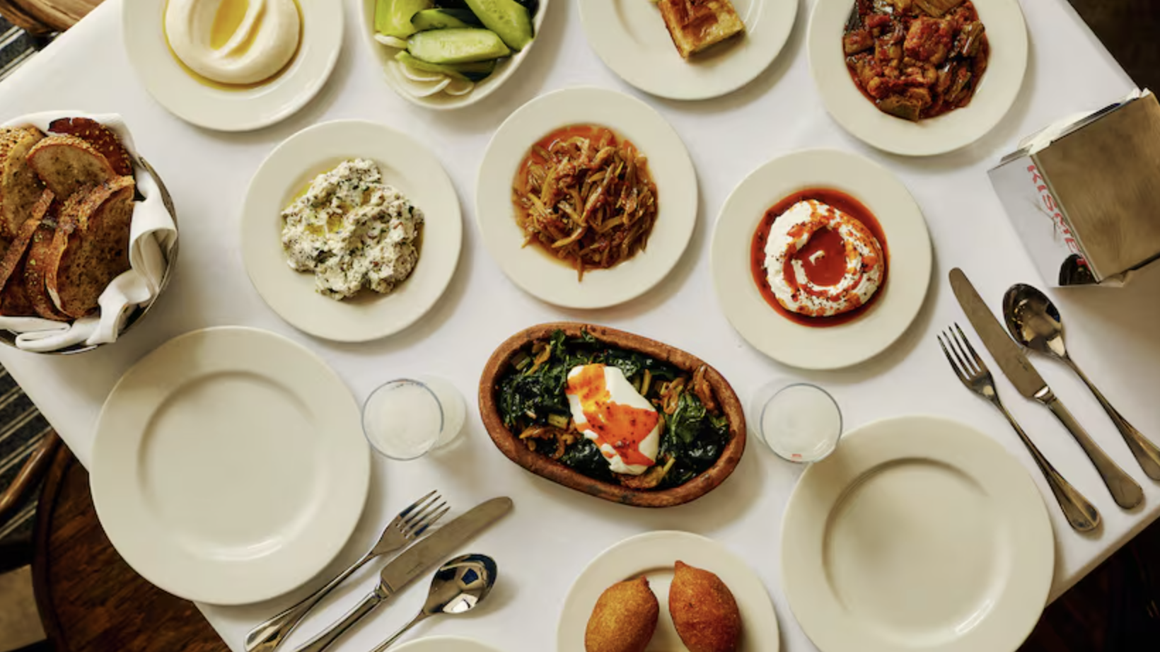 an image of nine different small plate style dishes on a white tablecoth covered table at Kismet, with a basket of bread in the corner