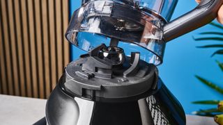 a black nutribullet with a big blending component is photographed in front of a blue backdrop, showing its plastic pitcher jug, silver blades, and tamper