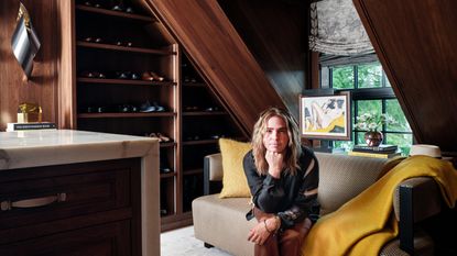 A young blonde man with long hair, dressed in a dark top and brown trousers, sits with his head resting on his first on a beige sculptural couch in a wood-clad room.