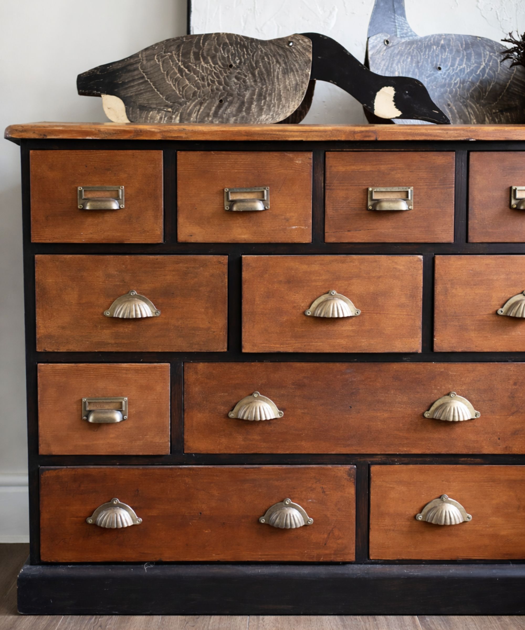 Vintage wooden dresser with brass pulls and cup handles, topped by carved goose decoys against a textured wall, styled in warm, natural light.