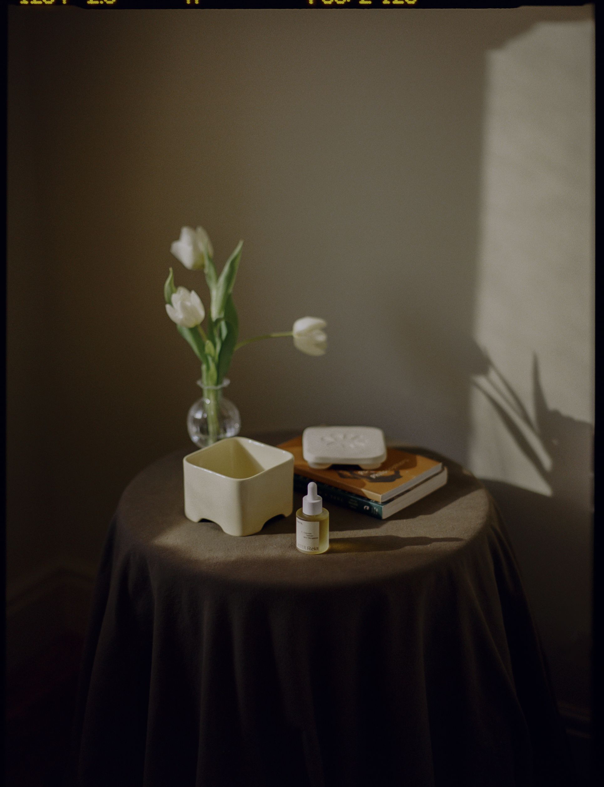 A room lit by a sunset boasts a home fragrance diffuser in the form of a pale yellow box, with a bottle of perfume, a vase of flowers, books, and other collectibles like candles and home accessories.
