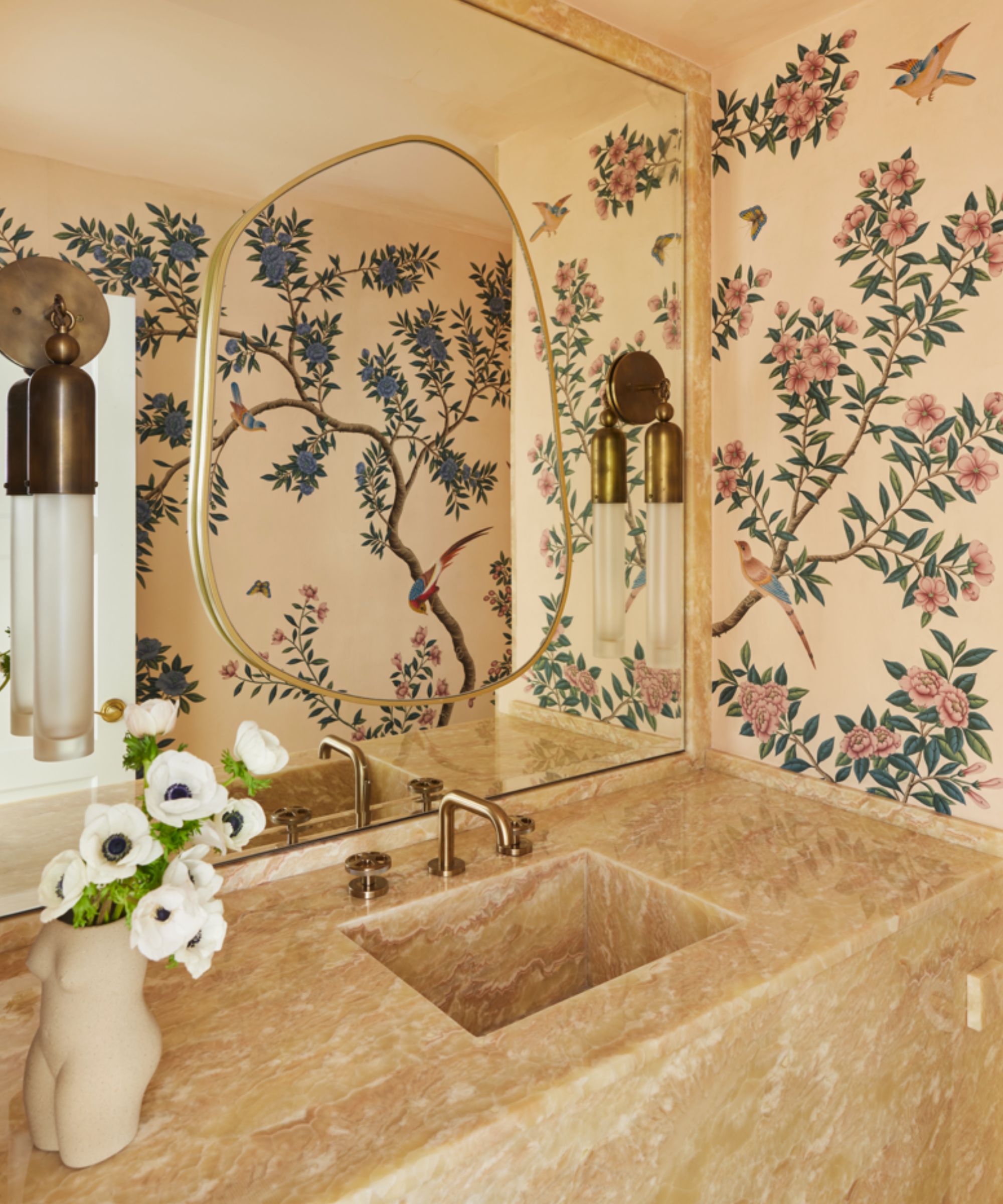 Corner of powder room with block stone sink, floral wallpaper, large wall to wall mirror with wall lights on top, vase of flowers to the bottom left