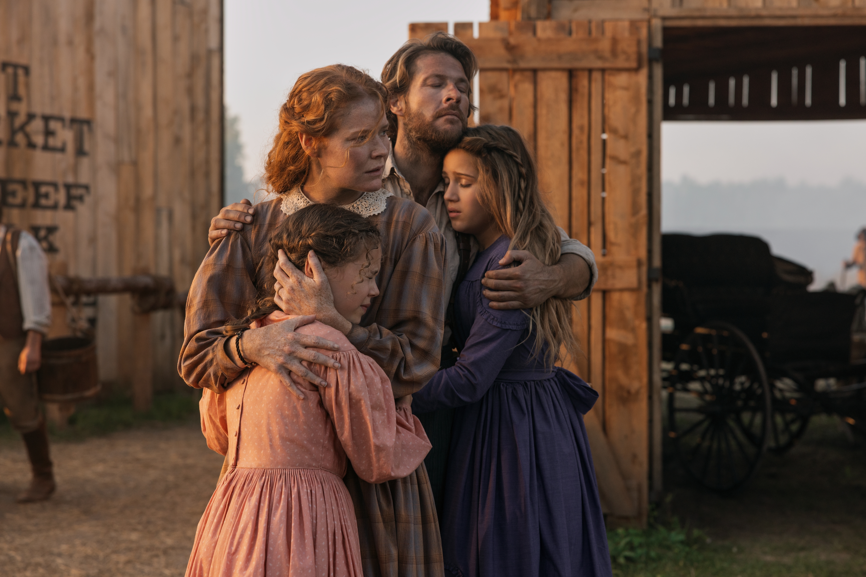 Alice Halsey as Laura Ingalls, Crosby Fitzgerald as Caroline Ingalls, Luke Bracey as Charles Ingalls, Skywalker Hughes as Mary Ingalls hug outdoors in front of wooden structures in 'Little House on the Prairie.'
