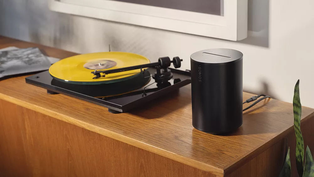 The Sonos Era 100 speaker pictured on a wooden surface next to a record player.