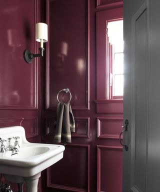 A powder room painted in Benjamin Moore's Carter Plum CW-355, a deep burgundy red, with traditional wall paneling, a white pedestal sink, silver hardware, and a black wall light finished with a white linen lamp shade