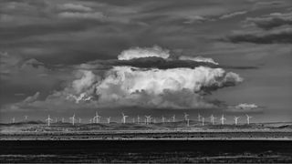 A monochrome image of a distant wind farm with large cloud looming above