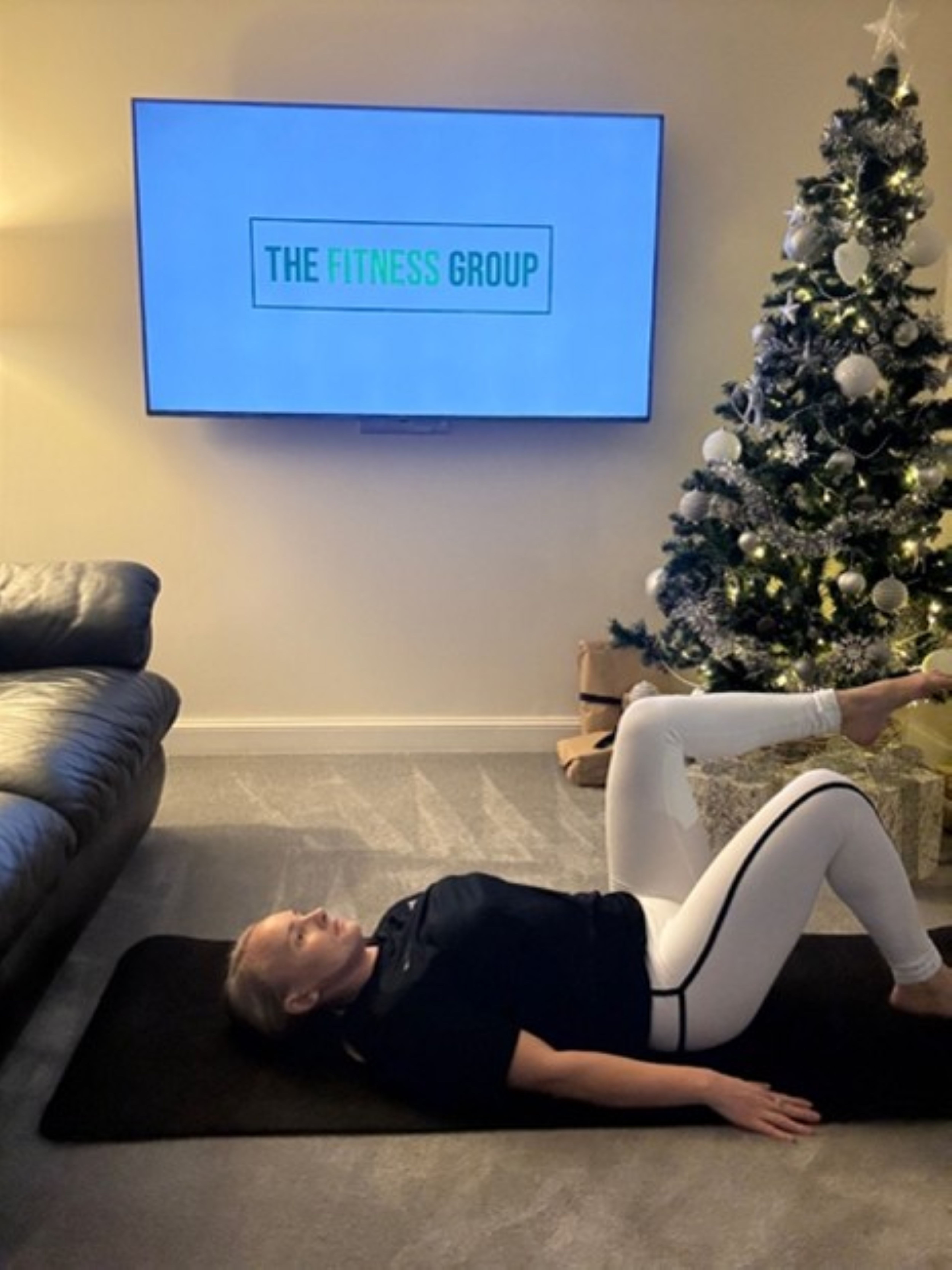 Woman demonstrates a Pilates exercise between a sofa and a Christmas tree. A mounted TV in the background reads "The Fitness Group"