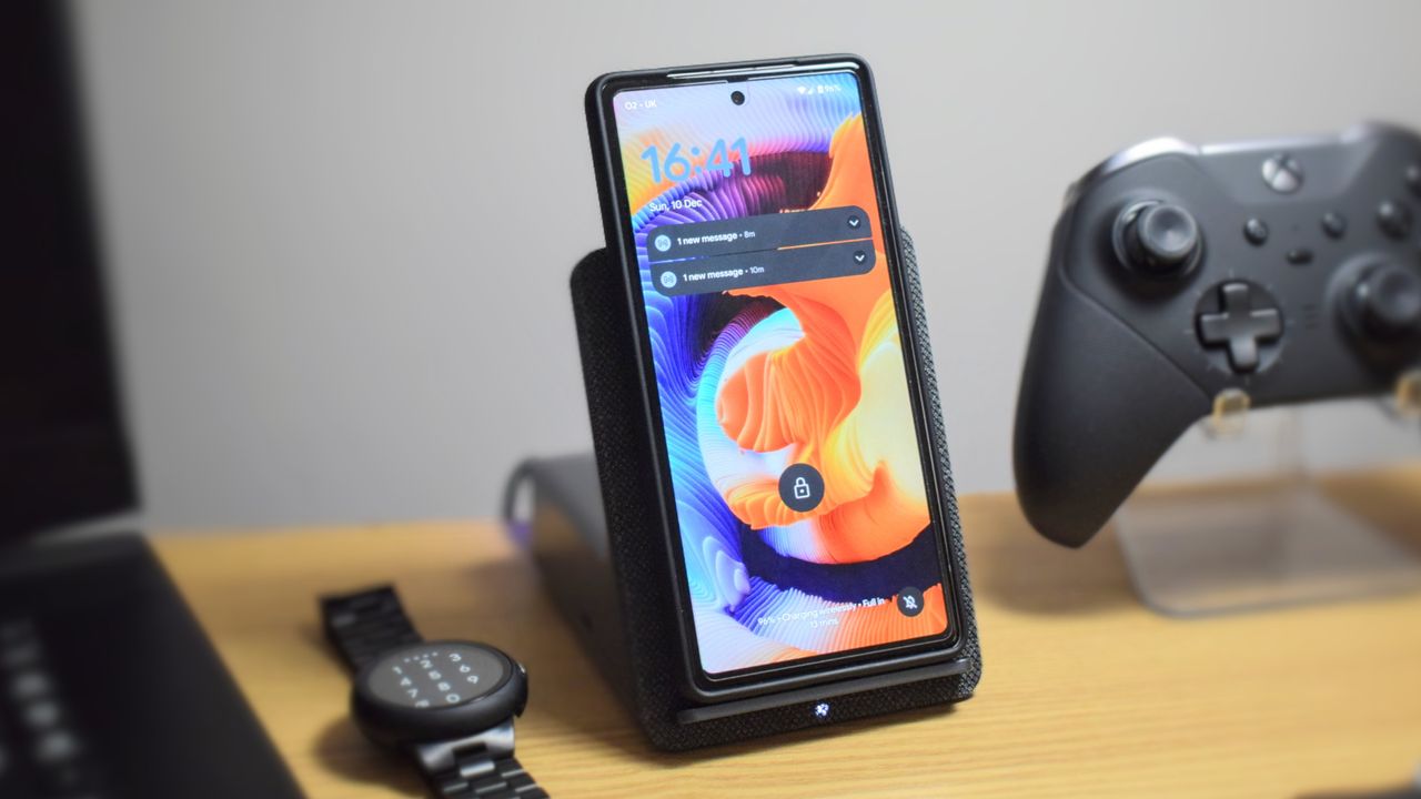 Photo of the Dell Dual Charge Dock with QI wireless charging docking station, showing a Google Pixel 7a in the wireless charging cradle. A Pixel Watch and Xbox Elite Wireless Controller Series 2 are next to the dock.