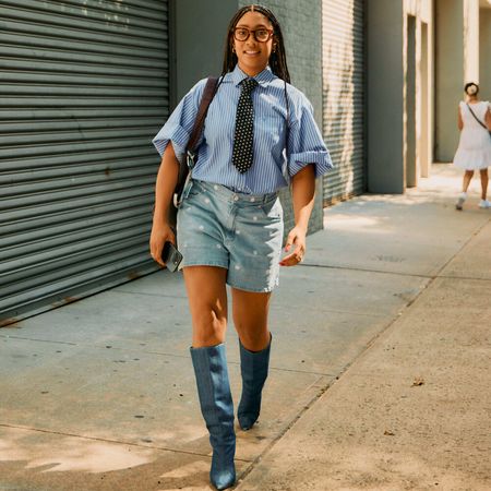 Woman wears short sleeve blue-striped button down with a tie, mid-length denim shorts, and tall denim boots while walking. 