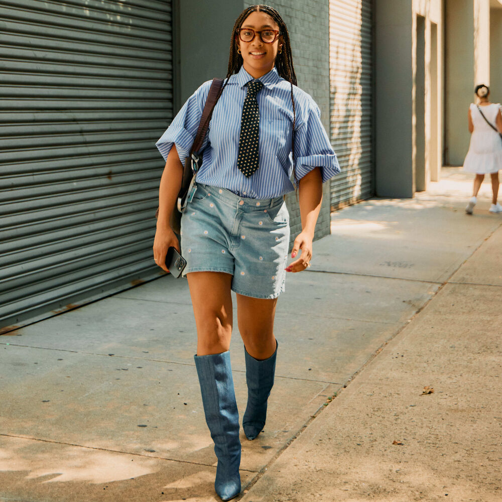Woman wears short sleeve blue-striped button down with a tie, mid-length denim shorts, and tall denim boots while walking. 