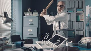 Angry businessman destroying his desk and laptop with a baseball bat
